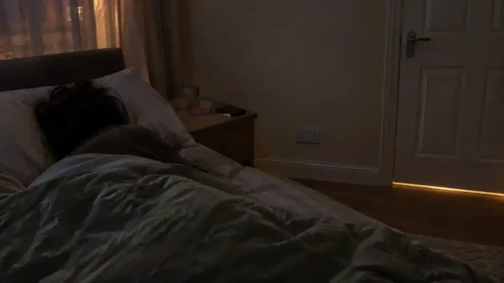 Person lying in bed in a dim UK rental bedroom with hallway light and noise leaking under the door