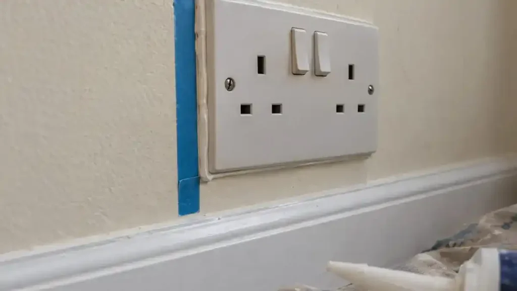 Sealing the gap around a UK double socket with acoustic sealant and masking tape to soundproof a thin rental wall