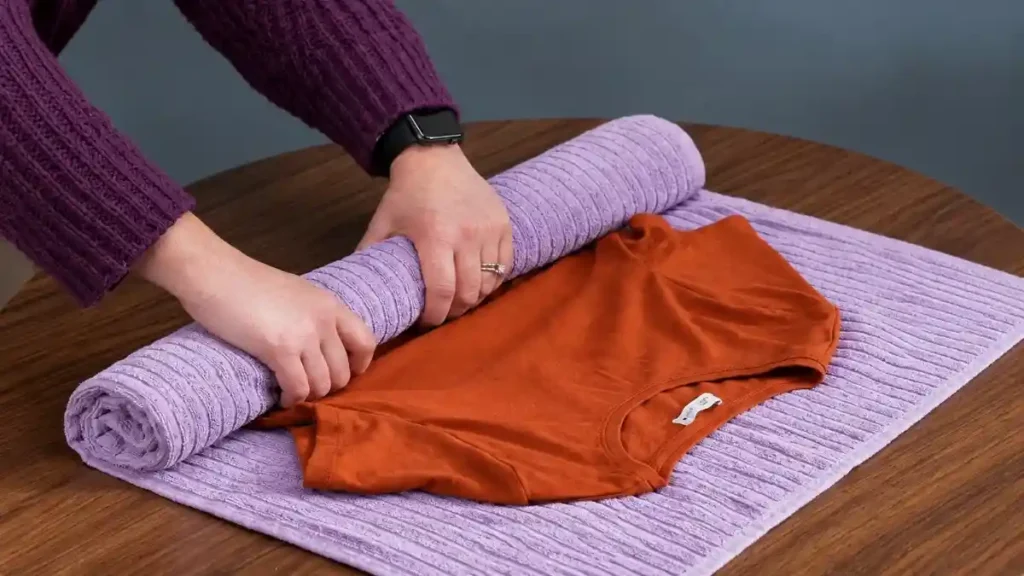 Rolling a wet orange t-shirt inside a dry lilac towel and pressing down to squeeze out water before hanging it to dry